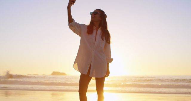 Woman capturing a moment on her phone at the beach during sunset. Ideal for travel promotions, casual lifestyle content, social media campaigns, or blogs about relaxation and vacations. Highlights the serene and beautiful backdrop of the ocean during the golden hour.