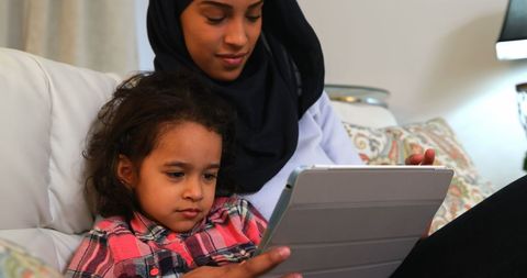 Mother in Hijab Reading with Daughter on Tablet in Cozy Home