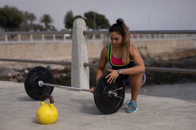 Front view of a strong Caucasian woman with long dark hair wearing sportswear exercising outdoors by the seaside on a sunny day, strength training preparing barbells, kettlebell next to het.