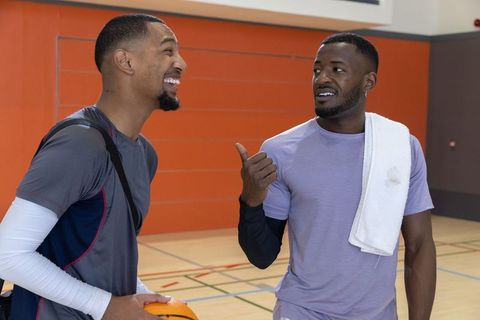 Two male friends laughing and talking on indoor basketball court after pickup game