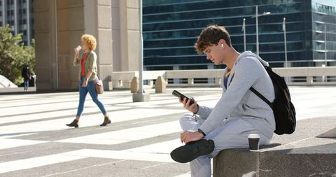 Young man sitting on city plaza ledge using smartphone with earbuds and backpack, casual urban lifes
