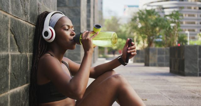 Young African American woman is sitting against a wall outdoors, drinking water after a workout. She is wearing wireless headphones and checking her smartphone, suggesting she is listening to music or tracking her workout. Ideal for content related to fitness, wellness, hydration, technology use in fitness, and active lifestyle.