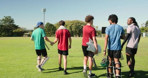 Five young cricket players walking across sunny outfield carrying bats, pads, helmet