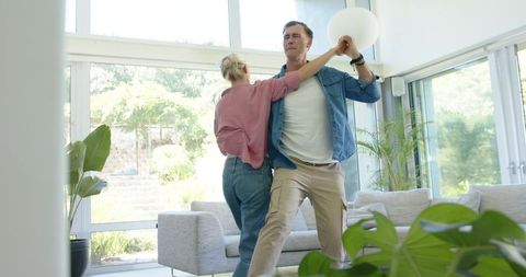 Middle-aged couple dancing in sunlit modern living room with pendant light and plants