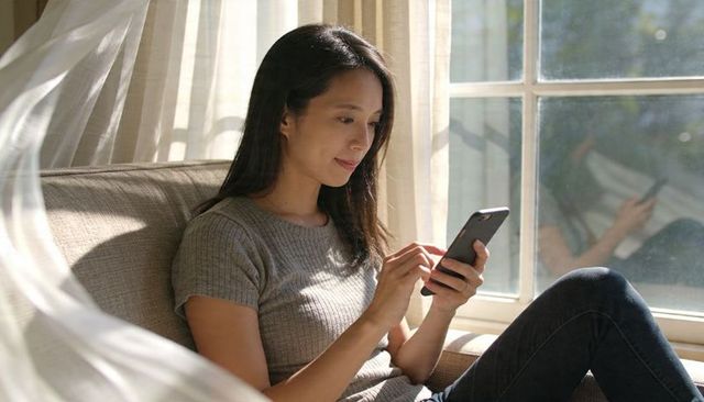 Woman relaxing on sofa scrolling smartphone by sunlit window with sheer curtains