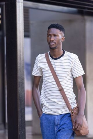 Young african man walking on urban sidewalk wearing striped tee, denim and leather bag