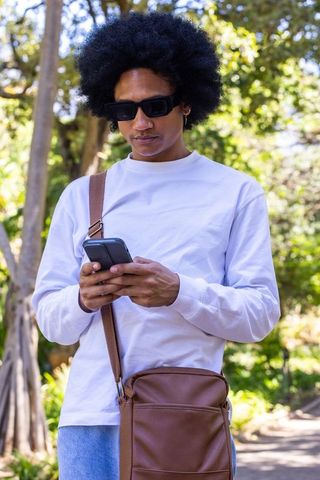 African american man checking smartphone outdoors with white shirt and brown crossbody bag