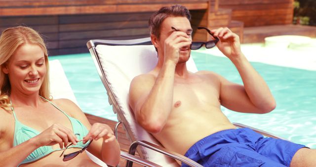 Couple is seated on lounge chairs near pool on a sunny day wearing swimwear while adjusting their sunglasses. Ideal for illustrating vacation lifestyle, leisure time, poolside relaxation, and summer activities.