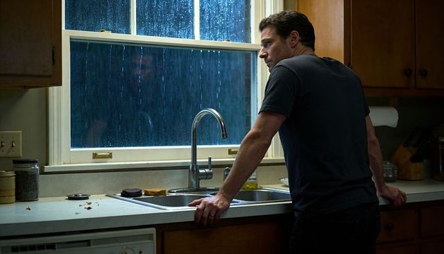 Man leaning at kitchen sink watching heavy rain through window, moody contemplative scene