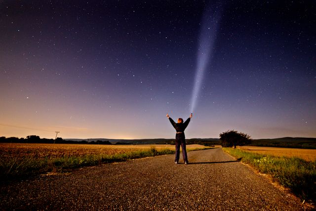 Young person standing on road in rural area with arms raised shining flashlight into the sky, creating a beam of light under starry night sky. Ideal for illustrating themes of exploration, independence, and adventure in nature. Perfect for use in travel blogs, outdoor gear promotions, and motivational content.