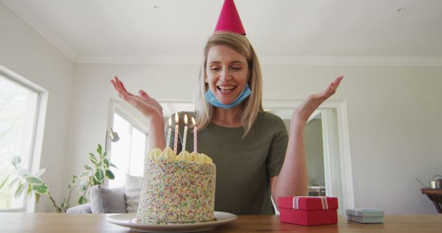 Woman enthusiastically celebrating birthday with a lit cake and presents at home. An animated individual wearing a mask hanging on her chin and a festive hat in a cheerful indoor environment. Perfect for use in ads, cards, social media posts, or blog articles highlighting birthday celebrations, joyful moments, home gatherings, and safety measures.