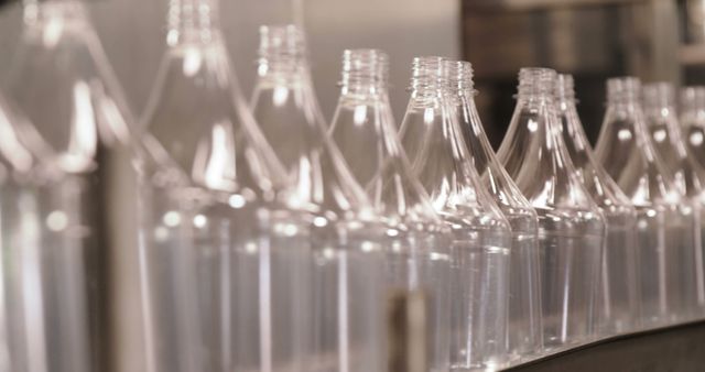 Clear plastic bottles are moving on an assembly line inside a factory. The focus is on their orderly arrangement and precision in the manufacturing process. Suitable for illustrating themes such as industrial production, beverage packaging, the plastic industry, and mechanical bottling processes.
