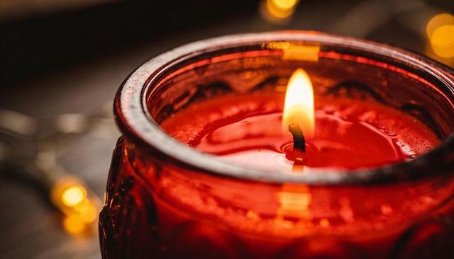 Glowing red jar candle casting warm reflections on dark tabletop, melting wax bokeh
