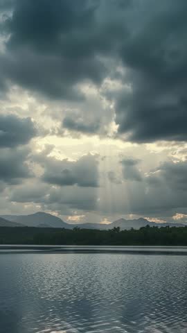 Vertical footage showing sunbeams piercing storm clouds and reflecting over mountain lake