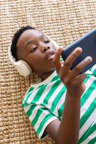 African american boy lying on jute rug wearing white headphones and using smartphone