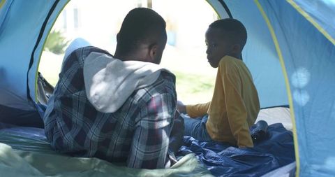 African american father and son bonding in backyard tent, sitting on sleeping bag