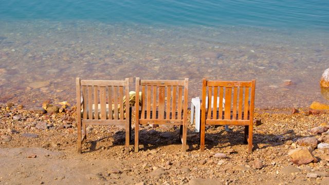Wooden chairs positioned on rocky shore with clear, calm water in background. Ideal for themes involving nature, relaxation, outdoor leisure, rustic furniture, and peaceful lakeside retreats. Useful for vacation advertisements, nature blogs, and lifestyle magazines.