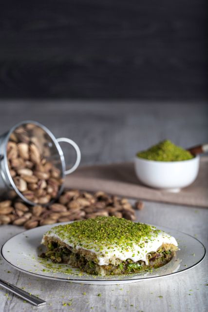 The image shows a delicious traditional Turkish dessert topped with crushed pistachios, elegantly plated. An ample amount of whole and ground pistachios is also visible, suggesting a rich pistachio use in the recipe. This image can be used for culinary blogs, recipe books or magazines, and food-related advertisements highlighting traditional delicacies.