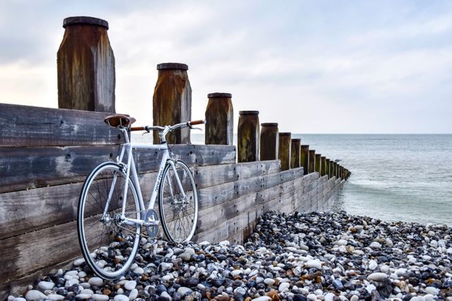 Vintage bicycle leaning on a wooden breakwater at a peaceful pebble beach. The calm ocean and overcast sky create a tranquil and serene atmosphere, perfect for themes of travel, summer, relaxation, and coastal living. Ideal for promoting outdoor activities, vacation spots, seaside resorts, and minimalist lifestyle content.