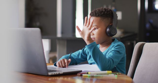 Young boy wearing headphones engaging in online class session, raising hand indicating participation. This can be used in content related to online education, home schooling, children's learning tools, remote learning technology.