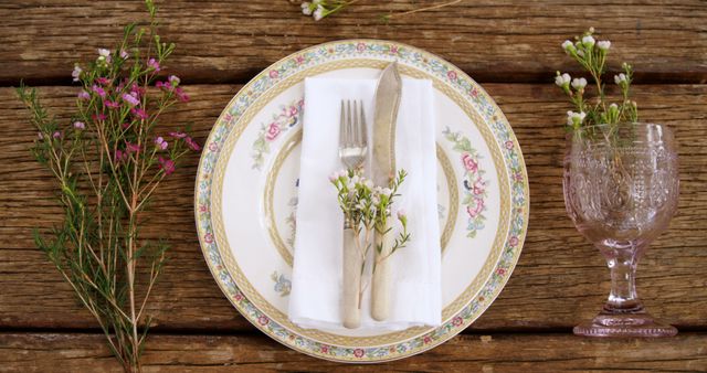 Elegant rustic table setting featuring vintage floral dinnerware arranged on a wooden table. Old utensils wrapped with floral bouquet add charm to the setting. Pink glass adds a delicate touch. Ideal for wedding, celebration, charming dining setup promotions, or lifestyle blogs focusing on decorating and dining ideas.
