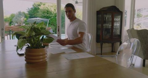 Young man reviewing paperwork at dining table with potted plant and patio view