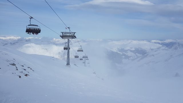 Ski lift moving over a snowy mountain landscape on a cloudy day provides a dynamic winter setting. Chairs dangle from the lift as they ascend the snowy slopes, suggesting travel and adventure in an outdoor winter sport environment. Use for travel blogs, winter sport promotions, or adventure articles illustrating cold weather activities and mountain scenery.