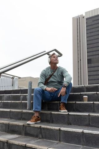 Middle-aged man listening on headphones sitting on urban steps with coffee and crossbody bag