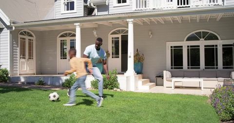 African american father coaching son playing soccer on front lawn of suburban home