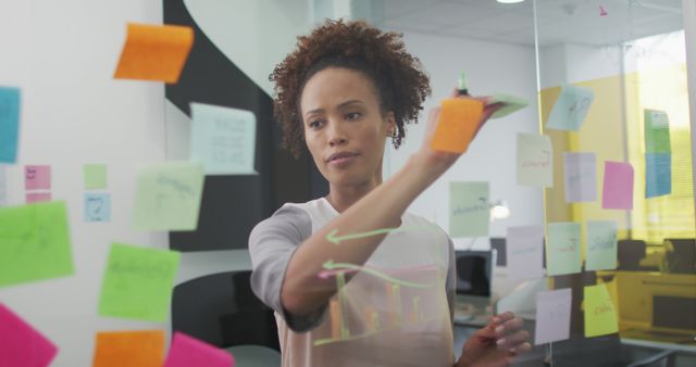 A young professional woman standing in front of a glass wall, applying sticky notes to organize ideas during a brainstorming session. Shows her in active thought, planning projects, or devising business strategies. Ideal for articles or advertisements about workplace productivity, creative thinking, business meetings, and female empowerment.