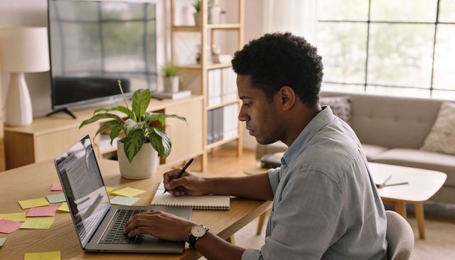 Focused man working from home on laptop while taking notes in spiral notebook with sticky notes