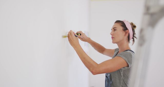 Woman carefully measuring white wall using a tape measure, wearing casual overalls and a pink headband. Ideal for illustrating home improvement projects, DIY tutorials, and precision in renovation tasks. Suitable for use in blogs, articles, and advertisements related to construction, home decor, and women's involvement in manual projects.