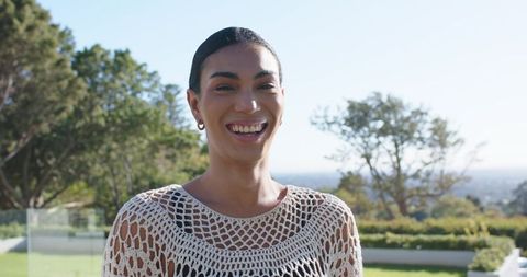 Smiling african woman standing on sunny terrace wearing white crochet top, hoop earrings