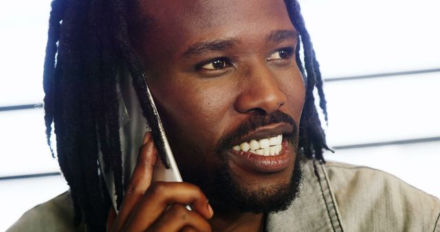 Young African American man with dreadlocks having a conversation on his cell phone indoors, exhibiting happiness through his smile. Ideal for advertisements, communication themes, technology products, customer service, and marketing materials aimed at casual, friendly interactions.