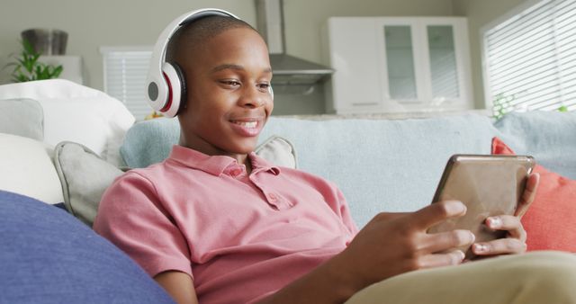 Image of happy african american boy in headphones using tablet on sofa. Childhood, spending time with technology at home concept.