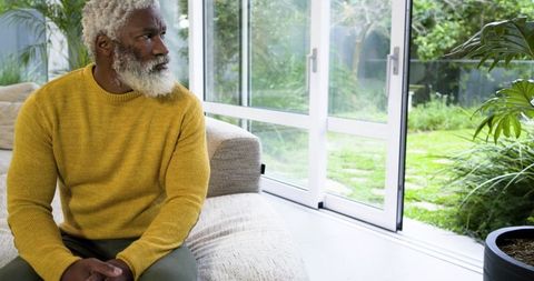 Senior African American man sitting on sofa in mustard sweater gazing through glass doors at garden