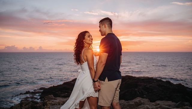 Romantic couple holding hands on rocky shore at sunset with flowing white dress
