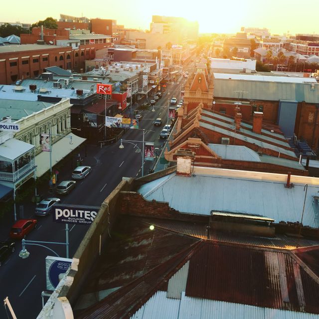 Image depicts an aerial view of a sunlit neighborhood street during sunset. Rooftops of various buildings, cityscape, and cars on the streets can be seen. Great for urban planning presentations, travel brochures, or articles showcasing city life.
