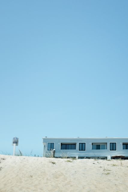 Modern beach house with large windows and a minimalistic design standing against a clear blue sky. Sand dune rises towards the front of the house. Ideal for use in real estate, travel, beach vacations, and summer lifestyle content.