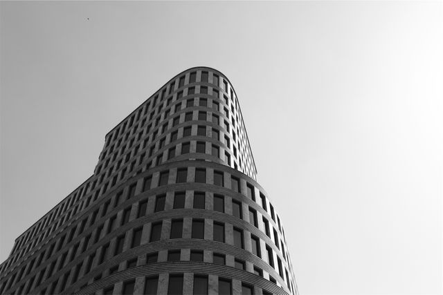 Impressive image of a modern curved skyscraper viewed from a low angle, highlighting its sleek design and architectural details against a clear sky. Useful for illustrating articles, blogs, or websites focusing on urban development, architecture, city planning, corporate structures, and minimalistic design ideals.
