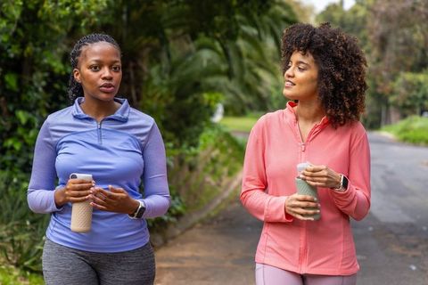 Two african american women walking and chatting on greenway in colorful activewear holding water bot