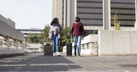 Diverse couple walking with rolling suitcases across urban pedestrian bridge, city travel