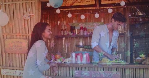 Smiling woman chatting with bartender slicing pineapple at bamboo tiki bar with lanterns and leis