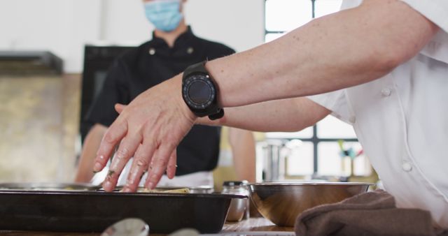 Chef wearing white uniform and black wristwatch prepares a dish in a professional kitchen. A colleague in a black chef's uniform and mask is present in the background. Useful for illustrating teamwork, culinary arts, professional food preparation, kitchen activities, or safety and hygiene practices in food service settings.