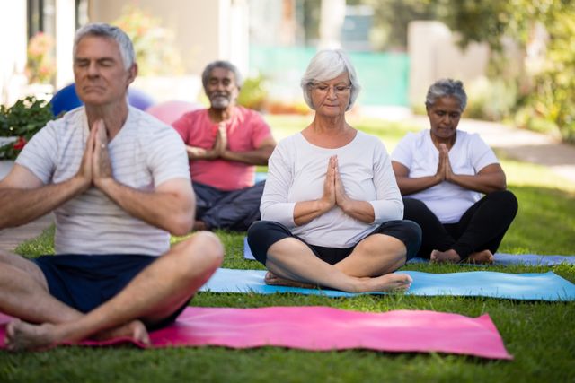Senior individuals are sitting on exercise mats in a park, meditating with closed eyes and hands in prayer position. This image can be used for promoting senior wellness programs, outdoor fitness activities, mindfulness and meditation classes, and healthy lifestyle campaigns for the elderly.