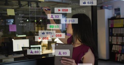 Woman writing on glass wall holding tablet annotating social analytics overlays in office