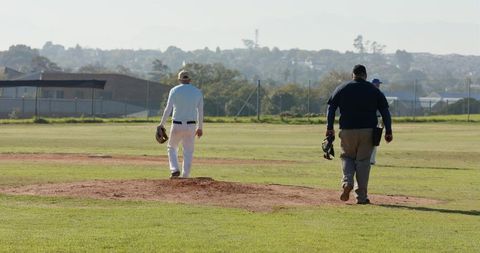 Three baseball players walking from pitcher mound carrying gloves on sunlit community field