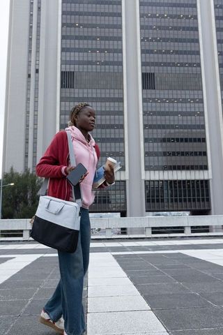African american woman walking downtown carrying coffee, phone, notebooks, tote bag