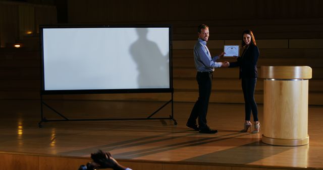 An individual is receiving an award on stage during a formal ceremony. They are shaking hands with the presenter, indicating a moment of recognition and achievement. A blank projector screen is in the background, highlighting the focus on the individuals and the awarded certificate. The image is well suited for illustrating themes like professional achievements, business events, recognition ceremonies, and public speaking engagements. It can be used in presentations, motivational materials, corporate communications, or event promotions.
