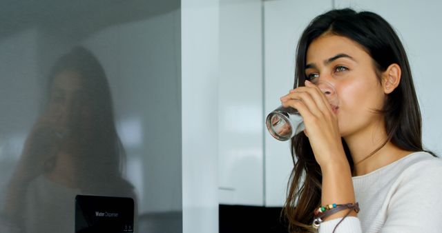 A young woman is drinking water in a modern kitchen, promoting a healthy lifestyle and hydration. This is perfect for websites or campaigns focused on healthy living, nutrition, and home life settings.